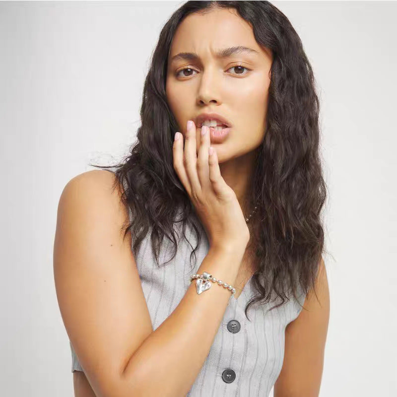 A woman with long, wavy dark hair poses in a light gray ribbed sleeveless top, wearing a silver charm bracelet on her wrist against a plain light gray background.
