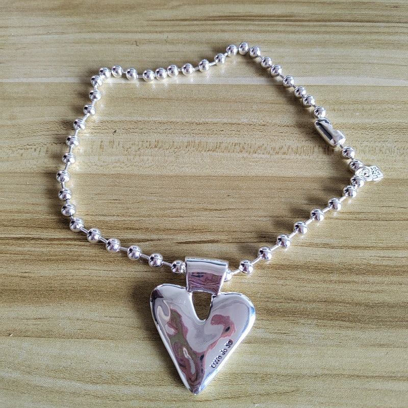 A silver heart-shaped pendant necklace with a beaded chain, displayed on a light wood grain surface.