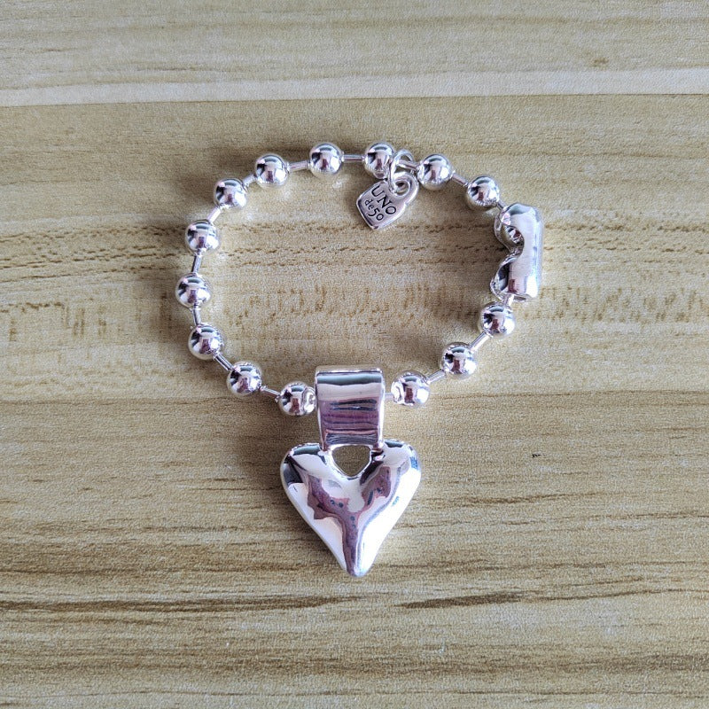 A silver beaded bracelet with a polished heart-shaped charm and a small heart tag engraved with 'LOVE', displayed on a light wood grain surface.
