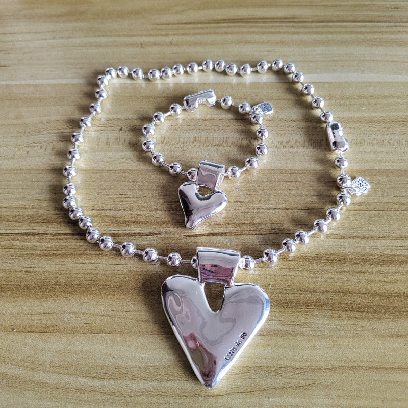 A sterling silver heart-themed jewelry set featuring a beaded bracelet and matching chain with a large polished heart pendant, displayed on a light wood surface.