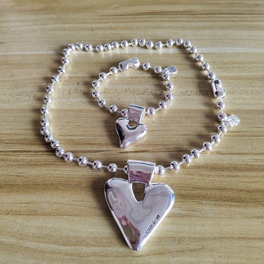 A sterling silver heart-themed jewelry set featuring a beaded bracelet and matching chain with a large polished heart pendant, displayed on a light wood surface.