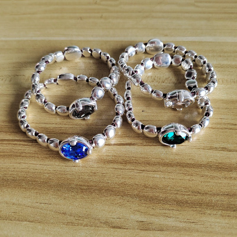 Three silver beaded stretch bracelets with oval gemstone settings, one blue, one green, and one clear, arranged on a light wood surface.