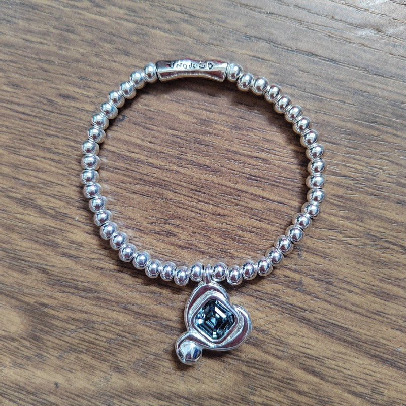 A sterling silver beaded bracelet with a heart-shaped charm featuring a dark blue emerald-cut gemstone, displayed on a wooden surface.