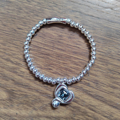A sterling silver beaded bracelet with a heart-shaped charm featuring a dark blue emerald-cut gemstone, displayed on a wooden surface.