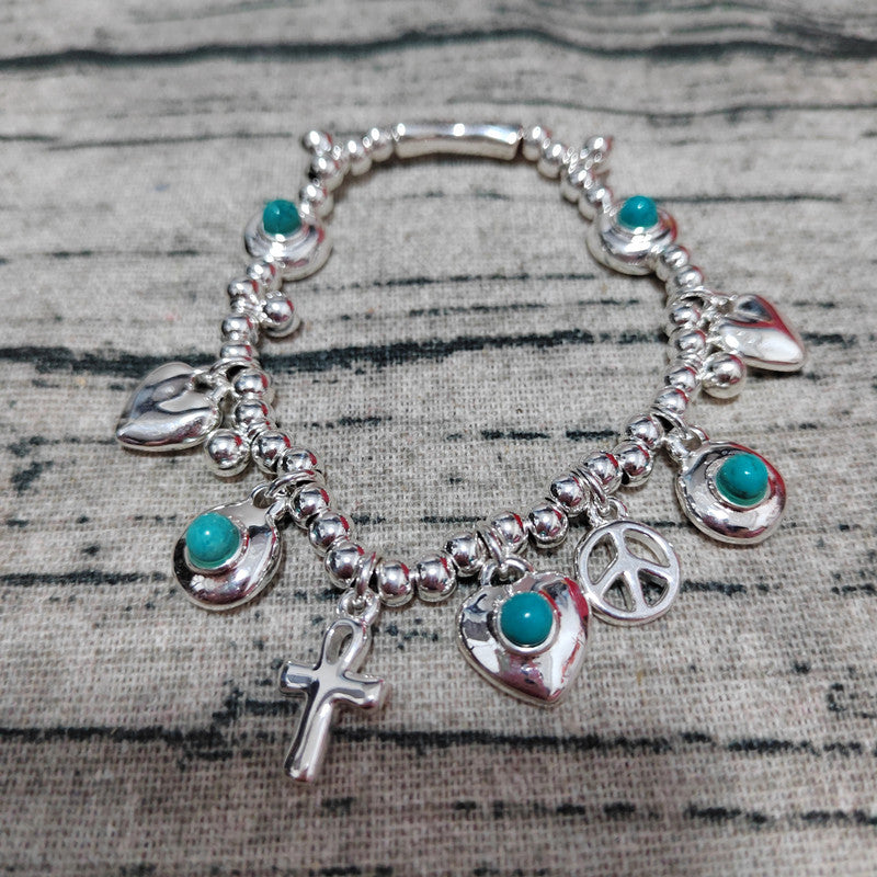 A silver charm bracelet with turquoise accents, featuring charms of a peace sign, cross, heart, crescent moon, and leaf, displayed on a textured gray fabric background.