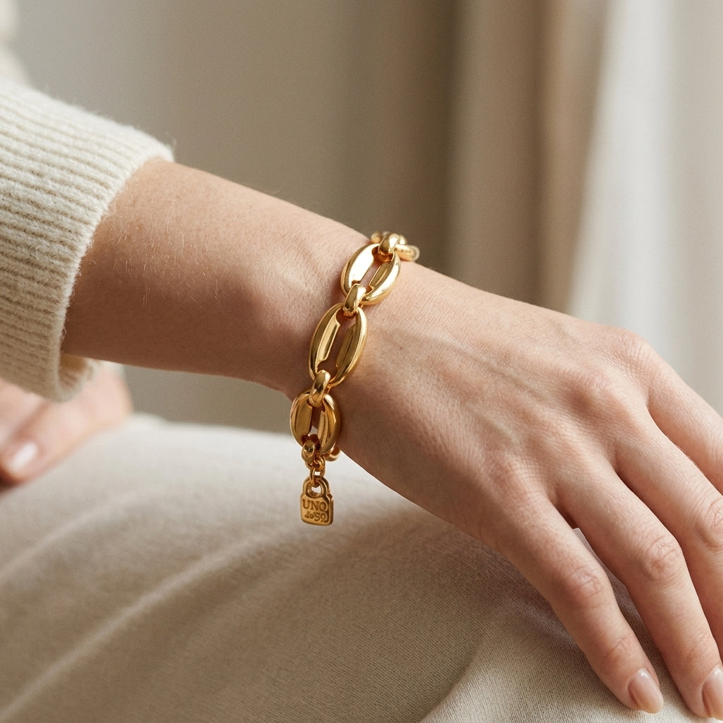 A close-up of a person's wrist wearing a polished gold chain bracelet with elongated oval links and a small hanging textured charm, paired with a cream ribbed sweater.