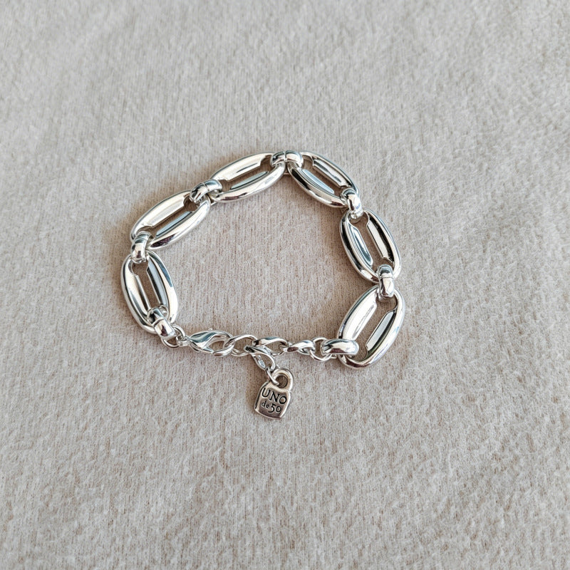 A silver chain bracelet with elongated oval links and a small hanging charm, photographed on a light beige textured fabric background.