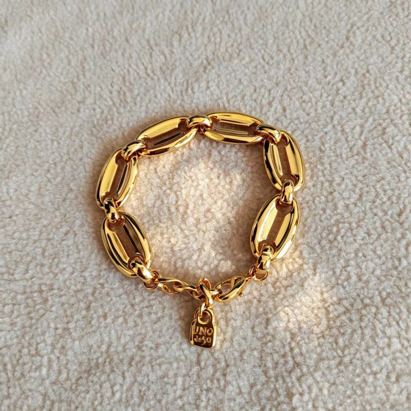 A gold chain bracelet with elongated oval links and a small hanging padlock charm, displayed on a textured cream fabric background.