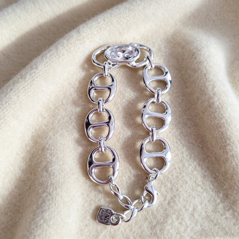 A silver-tone chain bracelet with interlocking oval links, a clear faceted crystal at the top, and a small hanging tag, displayed on a soft beige fabric background.