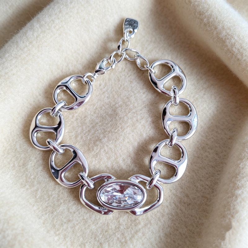A polished silver-tone link bracelet featuring a central oval clear crystal, displayed on a soft beige fabric background.
