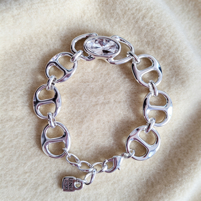 A silver-tone chain bracelet with interlocking oval links, a clear faceted crystal centerpiece, and a small hanging charm, displayed on a soft beige fabric background.