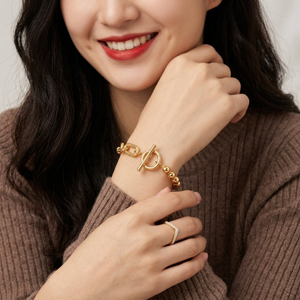 A close-up lifestyle shot of a woman wearing a gold chain bracelet with a toggle clasp on her wrist, paired with a matching gold ring on her finger. She is wearing a brown ribbed knit sweater against a soft, light gray background.