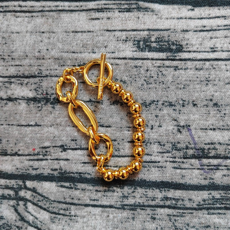 A gold-toned chain bracelet with a toggle clasp and a beaded stretch section, displayed on a textured gray fabric background.