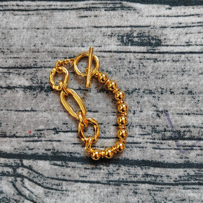 A gold-toned chain bracelet with a toggle clasp and a beaded stretch section, displayed on a textured gray fabric background.