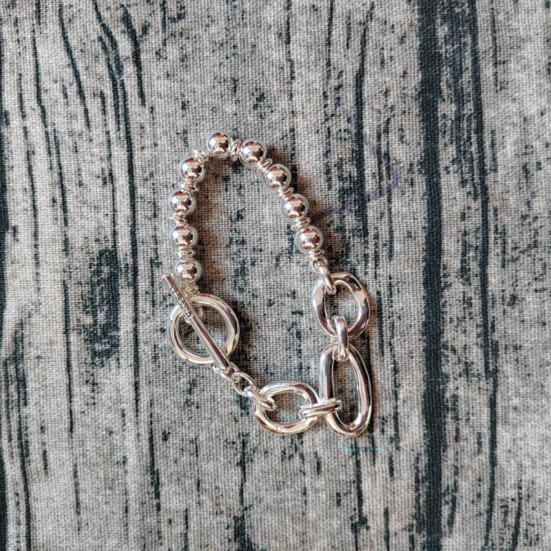 A silver-toned beaded bracelet with a toggle clasp and chain detail, laid flat on a gray wood-textured background.