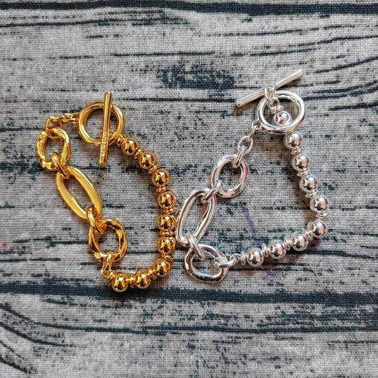 Two toggle clasp bracelets, one gold and one silver, displayed on a gray textured fabric background.