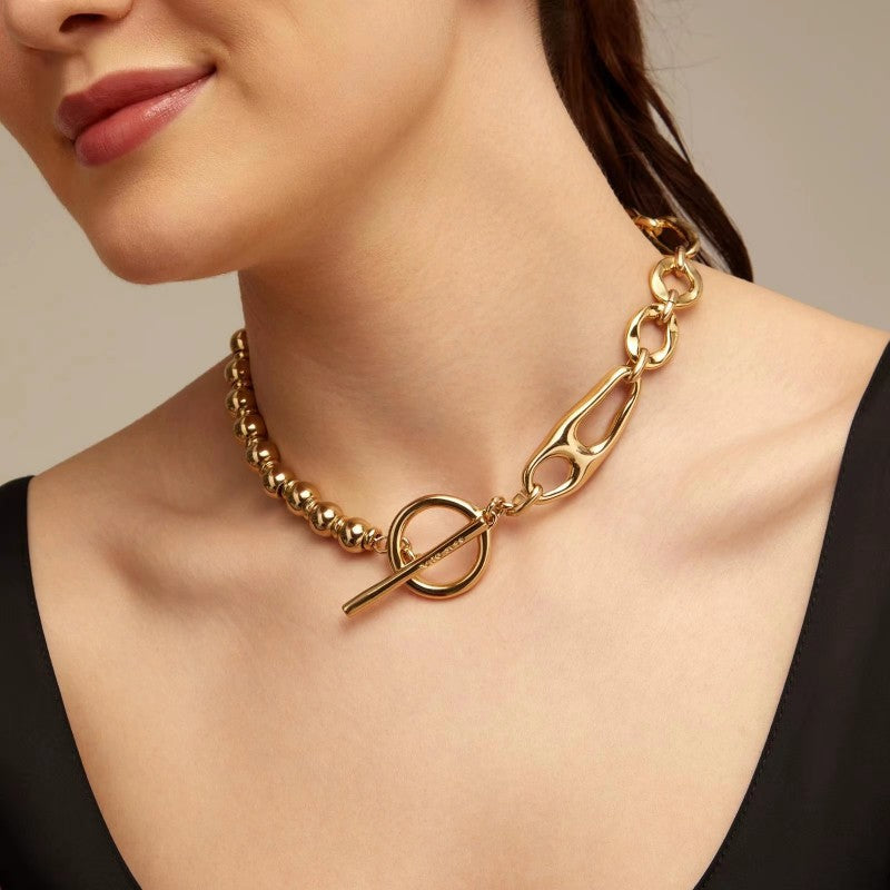 A close-up of a model wearing a gold-toned choker necklace with a beaded section, a large ring, and a toggle clasp, paired with a black top.
