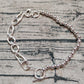 A silver-toned bracelet with a mixed link and bead design, featuring a lobster clasp, safety chain, and toggle clasp, displayed on a textured gray fabric background.