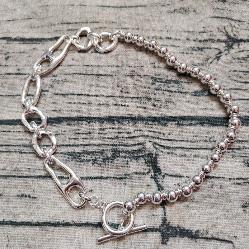 A silver-toned bracelet with a mixed link and bead design, featuring a lobster clasp, safety chain, and toggle clasp, displayed on a textured gray fabric background.