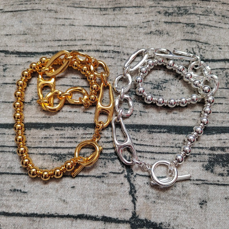 Two bracelets, one gold-toned and one silver-toned, displayed side by side on a textured gray fabric background. Each bracelet features a combination of a beaded chain and a chunky link chain with a toggle clasp.