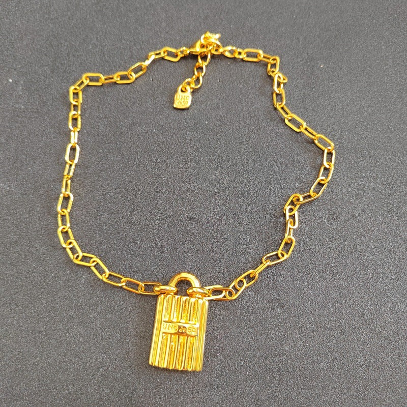 A gold-toned paperclip chain necklace with a small hanging padlock-shaped charm, displayed on a dark gray textured background.