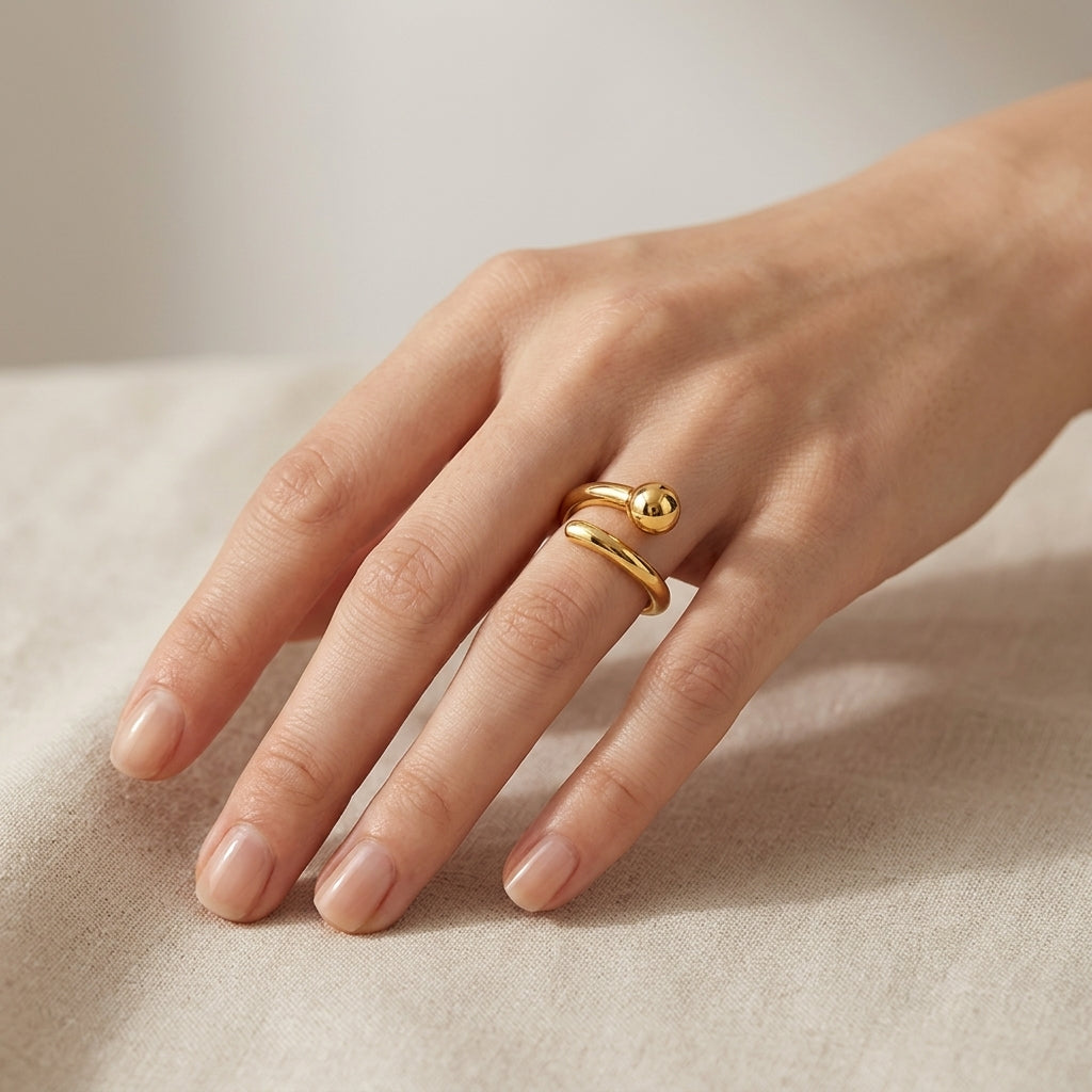 A close-up of a hand wearing a delicate gold ring with two curved bands and a small polished bead, resting on a soft beige fabric surface.