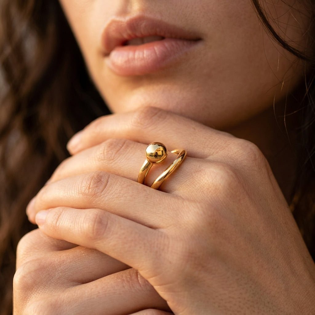 A close-up of a model's hand wearing a delicate gold ring with a polished spherical bead on a thin wrap-style band, set against a soft-focus background of her face and hair.
