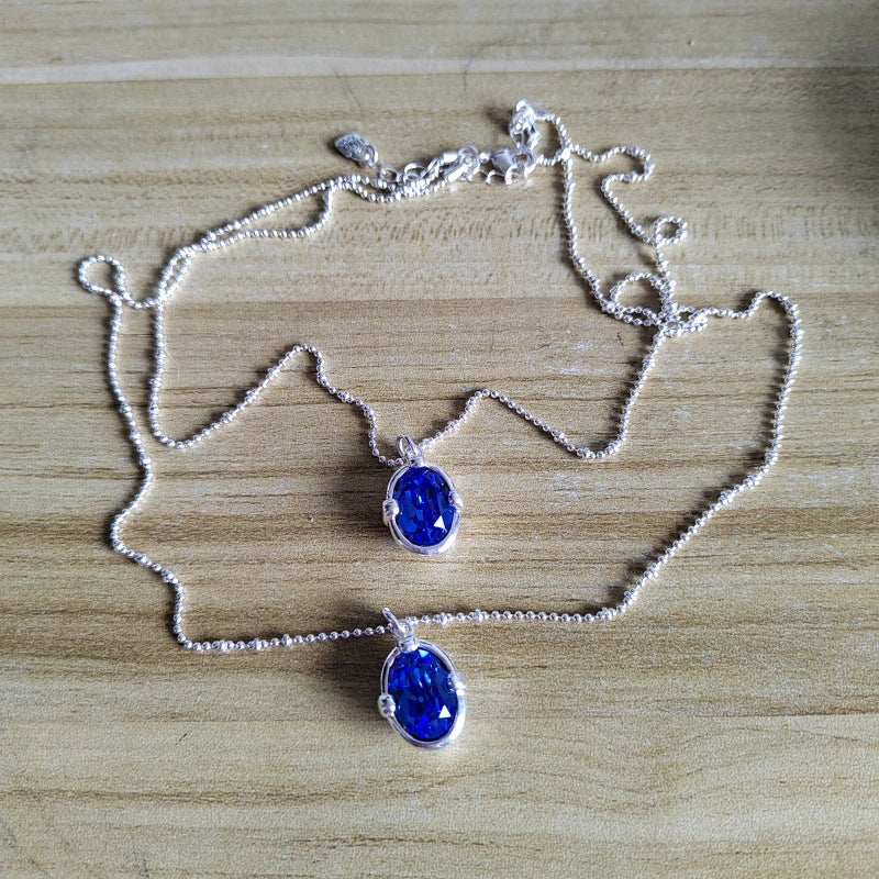 Two silver beaded chain necklaces with oval blue gemstone pendants, displayed on a light wood grain surface.