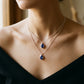 A close-up of a model wearing a double-layered necklace with two silver-tone pendants featuring deep blue teardrop gemstones, styled with a black off-the-shoulder top.