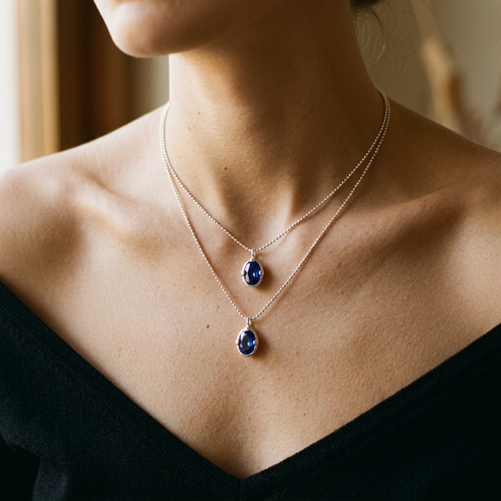 A close-up of a model wearing a double-layered necklace with two silver-tone pendants featuring deep blue teardrop gemstones, styled with a black off-the-shoulder top.