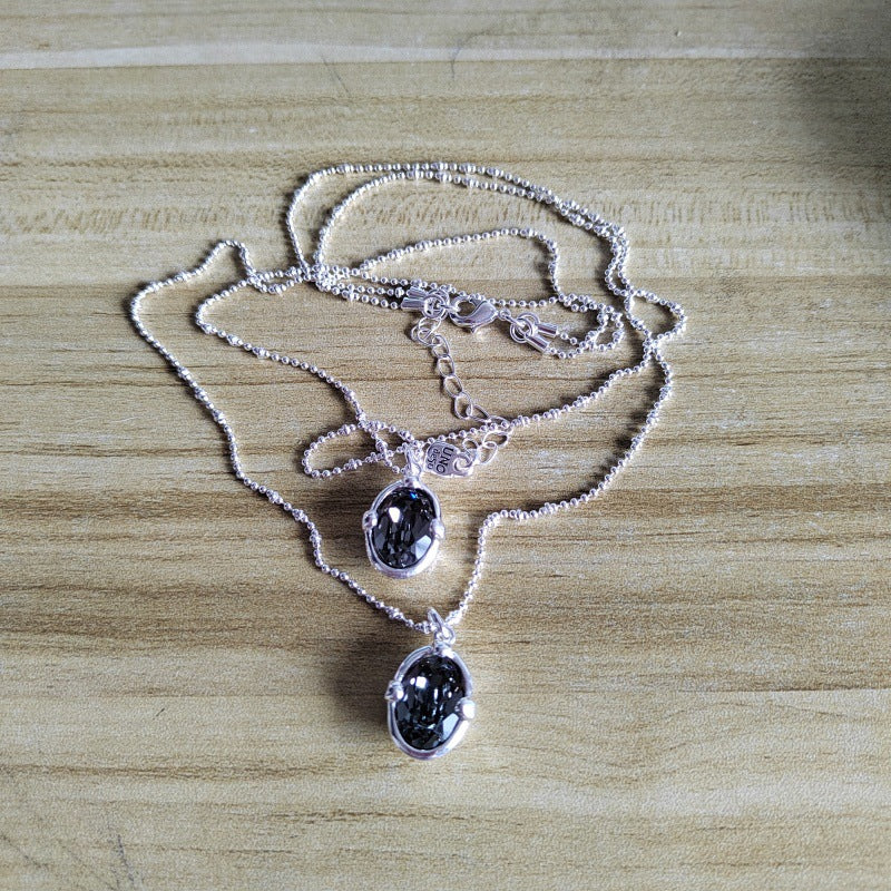 Two silver beaded chain necklaces with oval black gemstone pendants, displayed on a light wood grain surface.