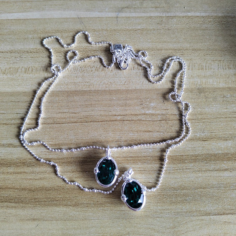 A silver beaded chain necklace with two dangling teardrop-shaped dark green iridescent gemstone pendants, displayed on a light wood grain surface.