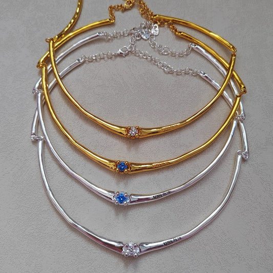 Three curved bangle necklaces stacked on a neutral gray background, featuring gold, silver, and mixed gold/silver bands with small blue and clear crystal accents.