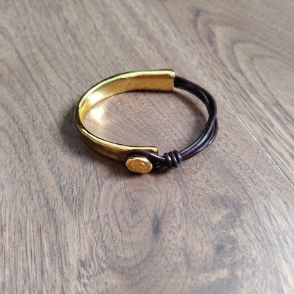 A gold-toned metal cuff bracelet with a dark brown cord wrap and a small round gold charm, displayed on a wooden surface.