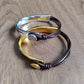 Three metal bangles stacked together on a wooden surface: a polished silver-tone bangle, a gold-tone bangle, and a dark brown cord bangle with a small round gold charm.