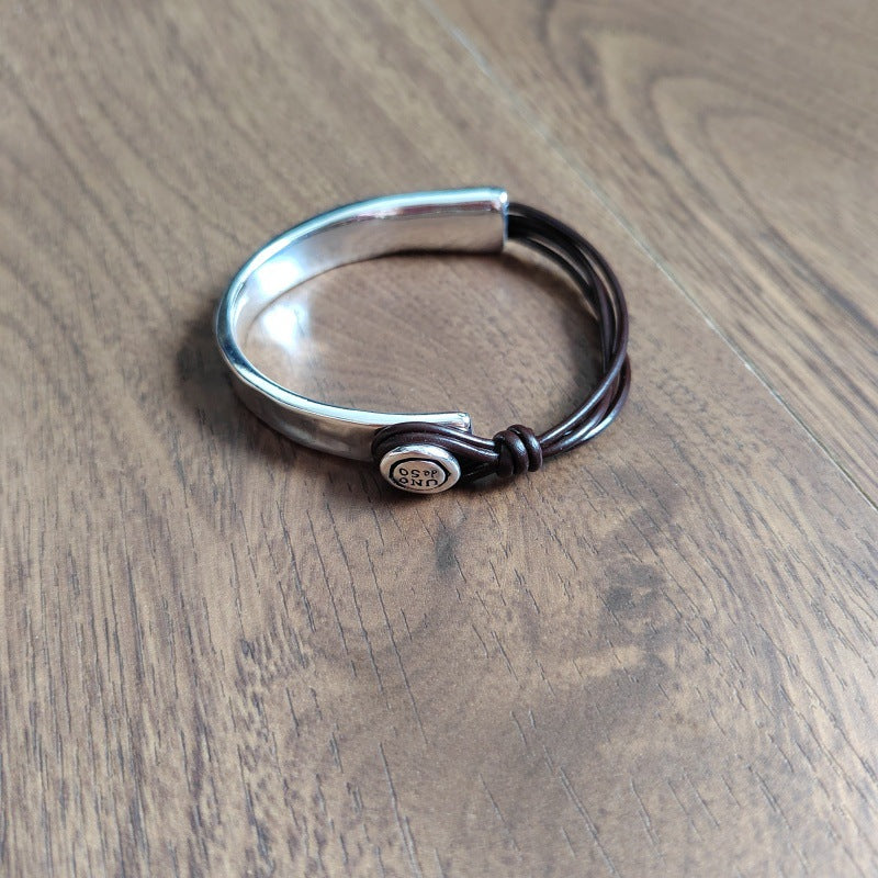 A silver-toned metal cuff bracelet with a dark brown leather wrap and a round silver charm engraved with 'SILVER CRAFT', resting on a brown wooden surface.