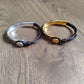 Two metal cuff bracelets with black cord wraps, displayed on a wooden surface. One bracelet is silver-toned with a round silver emblem, and the other is gold-toned with a round gold emblem.