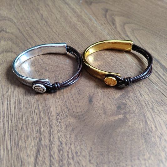 Two metal cuff bracelets with black cord wraps, displayed on a wooden surface. One bracelet is silver-toned with a round silver emblem, and the other is gold-toned with a round gold emblem.