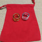 Two triple-band rings, one gold and one silver, displayed on a red drawstring pouch over a beige burlap background.