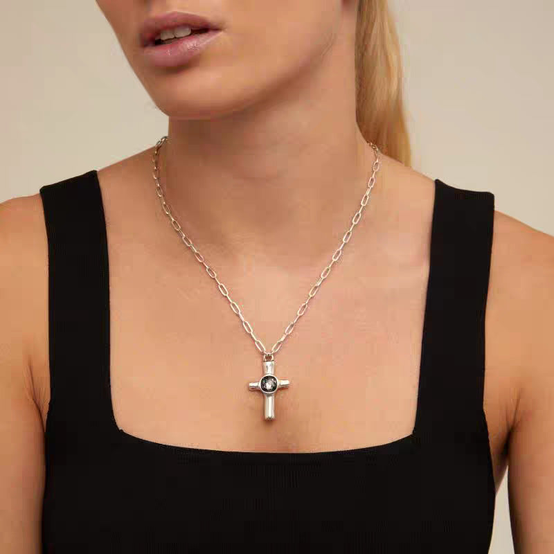 A close-up of a model wearing a silver cross pendant necklace with a black enamel center, styled with a black ribbed tank top against a warm beige background.