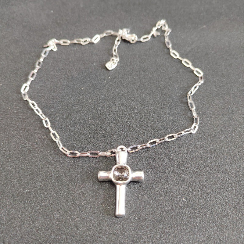 A silver chain necklace with a cross pendant featuring a clear round gemstone in the center, displayed on a dark gray textured background.