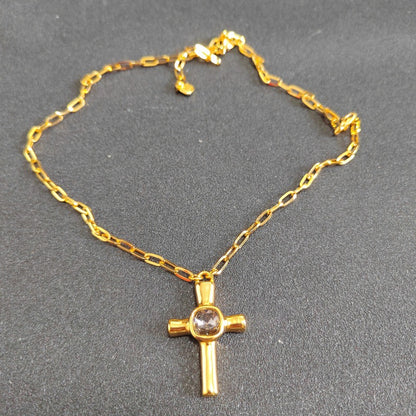 A gold cross pendant necklace with a clear round gemstone at the center, displayed on a textured dark gray background.