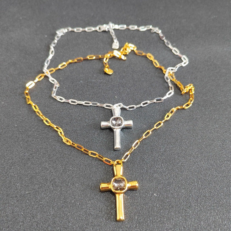 Two cross necklaces layered together on a dark gray textured background. The top necklace is silver with a silver cross and a clear round stone.