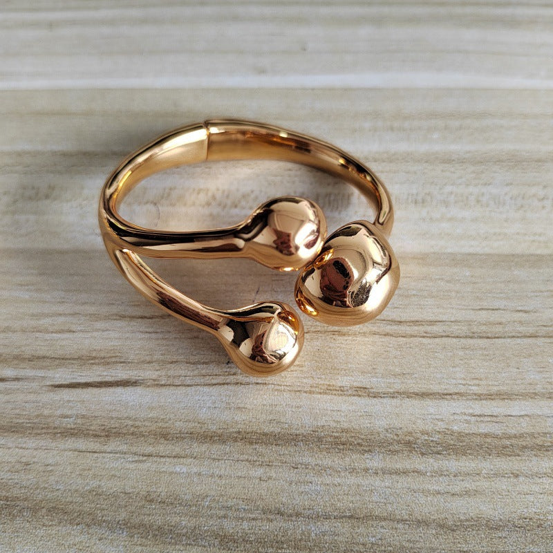 A polished rose gold metal stretch band bracelet with three rounded, organic-shaped protrusions, resting on a light wood grain surface.