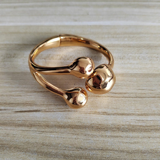 A polished rose gold metal stretch band bracelet with three rounded, organic-shaped protrusions, resting on a light wood grain surface.