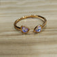 Gold-tone open cuff bracelet with two triangular iridescent purple-blue gemstones, resting on a light wood surface.