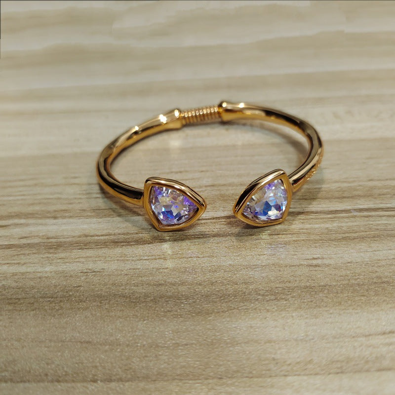 Gold-tone open cuff bracelet with two triangular iridescent purple-blue gemstones, resting on a light wood surface.