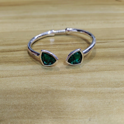 A silver-tone open cuff bracelet with two triangular green gemstone settings, displayed on a light wood surface.