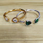 Two open cuff bracelets displayed on a light wood surface. One bracelet is gold-toned with a clear teardrop gemstone, and the other is silver-toned with a green teardrop gemstone.