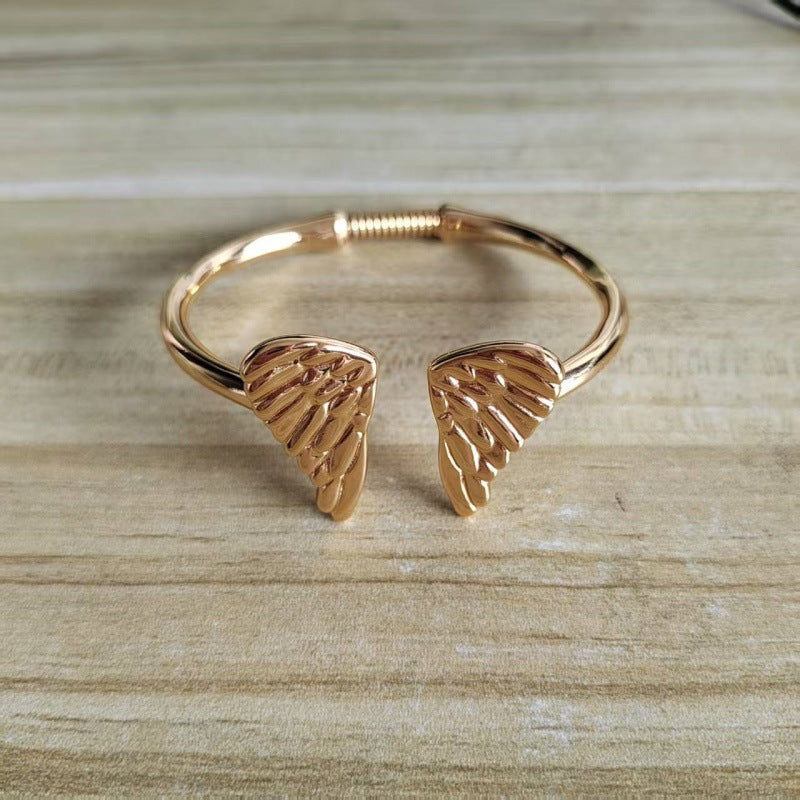 A rose gold cuff bracelet featuring two textured angel wing charms, displayed on a light wood grain surface.